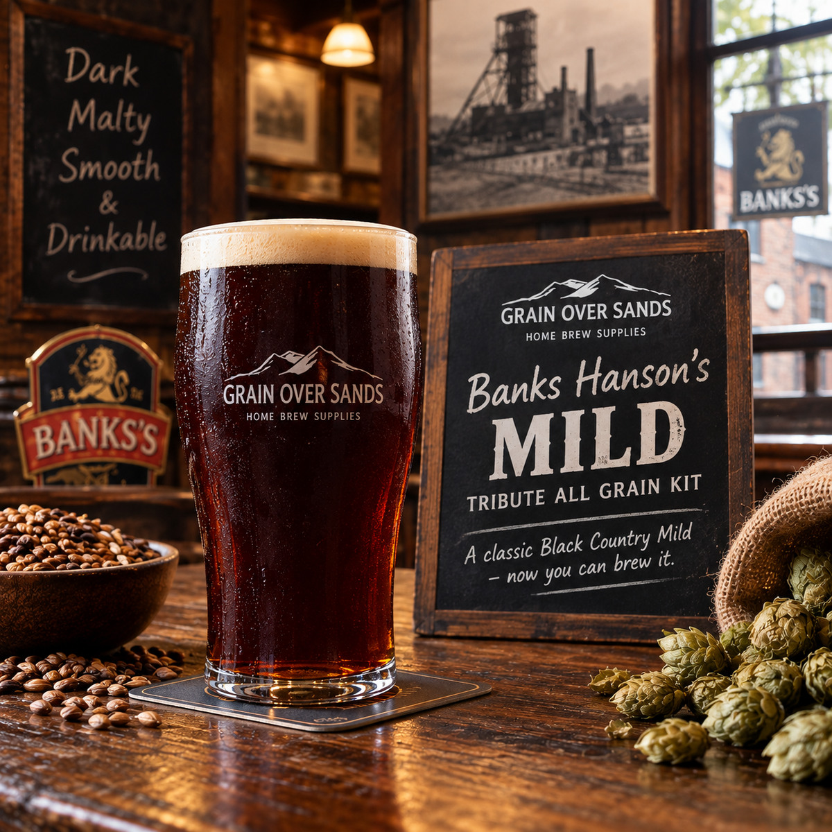 A frosted pint glass filled with dark beer stands on a coaster on a wooden bar, next to a chalkboard sign reading Grain Over Sands Banks Hanson's Mild Tribute All Grain Kit. Hop cones, a bowl of grain, and rustic brewery decor fill the background.