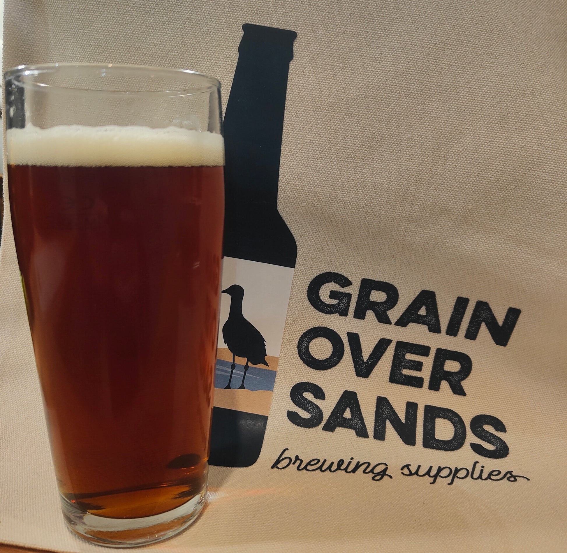 Glass of dark beer next to a 'Grain Over Sands brewing supplies' bag on a beige background
