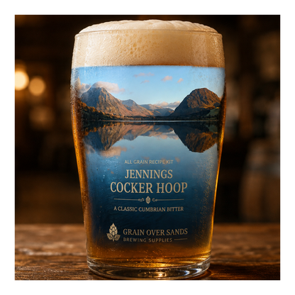 A pint glass of beer with a printed scenic label reading 'Jennings Cocker Hoop' and 'Grain Over Sands Brewing Supplies,' standing on a wooden table in a dimly lit pub setting.